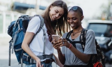 Two smiling women with backpacks look at a mobile phone.